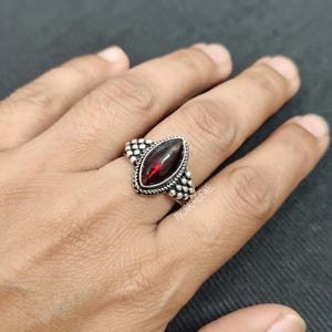 Op de afbeelding: Een zilveren ring met een donkerrode edelsteen in marquise-slijpvorm, gezet in een decoratief metalen frame met kraalachtige details langs de randen. De ringband heeft een textuur en de zetting is in het midden geplaatst.