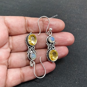 May include: Silver dangle earrings featuring oval yellow gemstones and smaller, round, opalescent stones. The earrings have a decorative design with intricate metalwork and a hook closure. The gemstones are set in a silver setting.