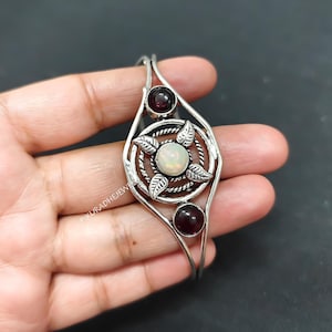 Op de afbeelding: Een zilveren manchetarmband met een centrale ronde witte opaal cabochon omringd door vier zilveren bladeren en een gedraaide draadring. Twee donkerrode granaat cabochons zijn boven en onder geplaatst. De tekst PIKURADHEJEWELRY is zichtbaar.