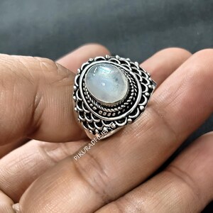 May include: A silver ring featuring a central oval moonstone cabochon. The stone is surrounded by a twisted rope border and a wide, decorative band with a repeating scalloped pattern. The text PIKURADHEJEWEL is visible on the metal surface.