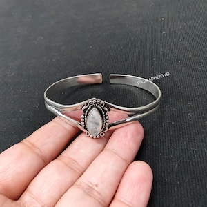 May include: A silver cuff bracelet featuring a central marquise-shaped moonstone cabochon set in a decorative beaded frame. The metal band splits into two thin arms that connect to the stone setting. The text PIKURADHEJEWEL is visible.