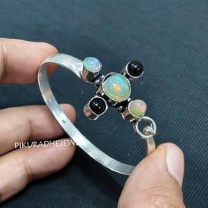 May include: A silver bracelet featuring a cross design with a central opal and four surrounding gemstones. The gemstones include black onyx and opals. The bracelet is held by a hand.