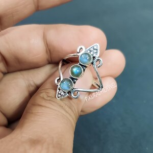 May include: A silver ring with three round, blue-green gemstones. The ring has a decorative triangular design with scrollwork and the text "PIKURADHEJEWEL". The gemstones are set in a row, and the ring is held by a hand.