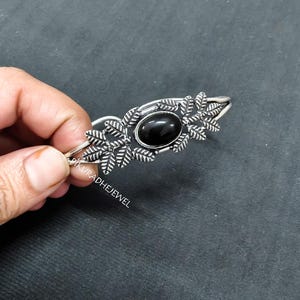 May include: Silver-toned cuff bracelet featuring an oval black gemstone center. The bracelet has leaf-like designs on either side of the gemstone. The jewelry is held against a dark gray background.