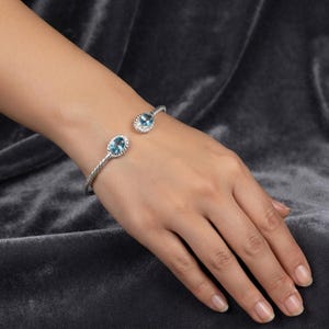 Könnte beinhalten: Ein silbernes Armband in Manschettenform mit einem gedrehten Metallband. Das offene Armband ist an beiden Enden mit einem ovalen blauen Edelstein verziert, der jeweils von einem kleinen, dekorativen silbernen Rahmen mit Perlenstruktur umgeben ist.