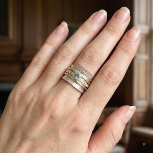 May include: A multi-band spinner ring featuring a central silver band with a light blue gemstone, flanked by two thin gold-tone bands and two wider silver bands with textured, etched patterns. The text PIKURADHEJEWEL is visible on the ring.