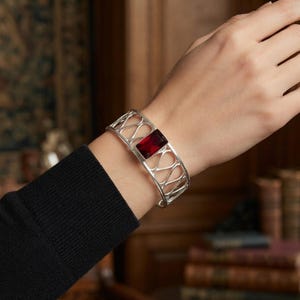 Puede incluir: Un brazalete de plata con un diseño de celosía calada y una sola piedra preciosa rectangular de color rojo intenso engastada en el centro. La banda de metal tiene un acabado pulido y rodea la muñeca con un patrón geométrico de bucles entrelazados.