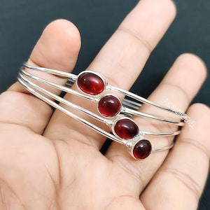 Könnte beinhalten: Ein Set aus vier silbernen Armreifen, jeder mit drei ovalen, tiefroten Edelsteinen. Die Armreifen sind gestapelt, wodurch das polierte Metall und das leuchtende Rot der Steine hervorgehoben werden. Der Schmuck wird auf einer Hand präsentiert.