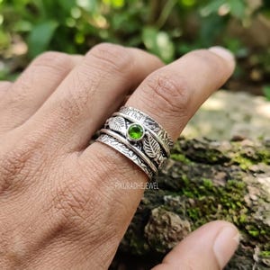 Könnte beinhalten: Ein silberner Spinner-Ring mit einem Mittelband, das einen kleinen, runden, grünen Edelstein und eingravierte Blattmotive aufweist. Der Ring hat eine texturierte, oxidierte Oberfläche mit mehreren drehbaren Bändern und dem Text PIKURADHEJEWEL.