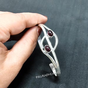 Könnte beinhalten: Ein silberfarbenes Armband im Manschettenstil mit einem geteilten Schaftdesign und zwei ovalen, dunkelroten Cabochon-Steinen in Metallfassungen. Die Metalloberfläche hat eine texturierte, matte Optik. Der Text PIKURADHEJEWEL ist unten in Weiß aufgedruckt.
