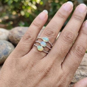 May include: A stack of three silver rings, each featuring a small, oval-shaped iridescent opal gemstone. The bands of the rings have a twisted, rope-like texture. The text PIKURADHEJEWEL is visible on the metal surface near the bottom gemstone.