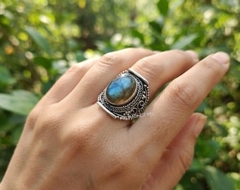 May include: A silver ring featuring an oval-shaped labradorite gemstone with blue iridescence. The metal band displays intricate decorative wirework and beaded detailing surrounding the central stone. The text PIKURADHEJEWEL is visible.