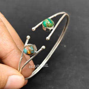 Könnte beinhalten: Ein silberfarbenes Armreif-Armband mit zwei runden, farbigen Steinen. Einer ist grün, der andere ist eine Mischung aus Türkis, Braun und Gold. Das Armband hat ein einzigartiges Design mit geschwungenen Linien und kleinen Kugelakzenten.