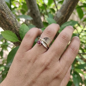 Può includere: Un anello con una piccola pietra preziosa rossa di forma ovale incastonata in argento, accentuata da una fascia in argento testurizzato che avvolge un sottile filo color oro. La superficie metallica presenta un motivo inciso e il testo PIKURADHEJEWEL.