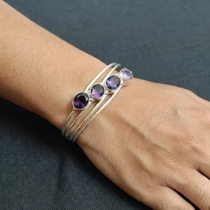May include: A silver cuff bracelet featuring three thin, parallel bands and four round, faceted purple amethyst gemstones set in a row along the top. The stones vary slightly in shade, ranging from deep violet to a lighter, translucent lavender hue.