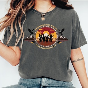 May include: Dark gray t-shirt with a circular graphic celebrating Indigenous Peoples' Day. The design features silhouettes of dancers, mountains, eagles, and the text "Honor Indigenous Wisdom" and "Indigenous Peoples' Day."
