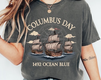 Könnte beinhalten: Dunkelgraues T-Shirt mit einer Grafik, die drei Segelschiffe auf dem Ozean zeigt, mit dem Text "Columbus Day" darüber und "1492 Ocean Blue" darunter. Das Design beinhaltet eine Windrose und Wolken.