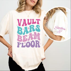 May include: Cream-colored t-shirt with the words "VAULT BARS BEAM FLOOR" in a retro font, in pink, teal, lavender, and purple. The sleeve has the name "Sophia" and two hearts in pink. The shirt is a relaxed fit.