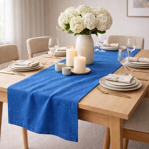 May include: A dining table set for a meal, featuring a vibrant blue table runner. The table is set with white plates, napkins, and gold-colored cutlery. A white vase holds a bouquet of white flowers, and candles add a warm ambiance.