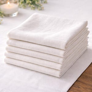 May include: A stack of six folded white linen napkins. The napkins are neatly arranged, showcasing their clean, crisp edges and soft texture. The image is well-lit, highlighting the bright white color of the fabric. The napkins are on a white surface.