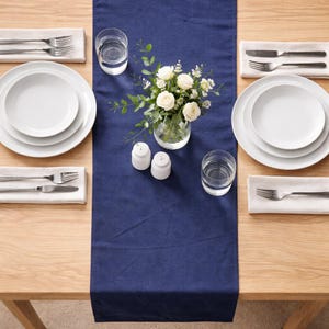 May include: A dining table setting with a navy blue table runner, white plates, silverware, and clear drinking glasses. A small vase of white roses and greenery sits in the center. Salt and pepper shakers are also present.