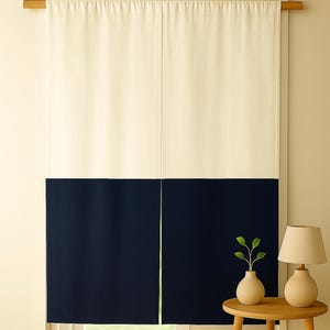 May include: A two-panel curtain with a cream-colored top and a navy blue bottom. The curtain hangs from a wooden rod above a window. A small wooden table holds a vase with greenery and a lamp.