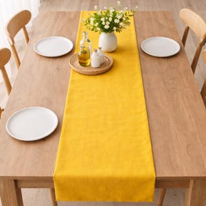 May include: A wooden dining table set with a bright yellow table runner. The table is set with white plates, wooden chairs, and a centerpiece of flowers in a white vase. A small tray holds bottles of oil and vinegar.