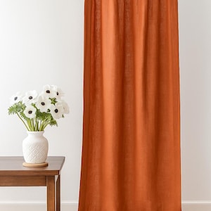 Puede incluir: Un panel de cortina de lino naranja quemado con construcción de lengüetas. La cortina cuelga de una barra de madera. Un jarrón blanco con flores blancas se encuentra sobre una mesa auxiliar de madera. La cortina es de color liso y tiene una apariencia texturizada.