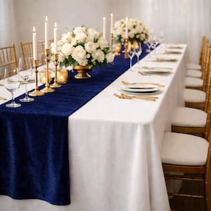 May include: A long dining table set for a formal event. The table is covered with a white tablecloth and a navy blue velvet runner. Gold chairs, candle holders, and cutlery complement the white floral arrangements.