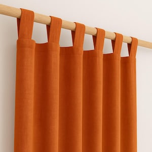 Puede incluir: Primer plano de cortinas naranjas con presillas colgadas de una barra de madera. Las cortinas son de color liso y tienen un aspecto texturizado. Las presillas son del mismo color que las cortinas. El fondo es una pared blanca lisa.