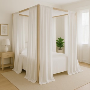 May include: A four-poster bed with a light wood frame and white, flowing curtains. The bed is made with white linens and is in a bright room. A small bedside table with a lamp and a framed picture are also visible.