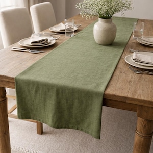 Puede incluir: Una mesa de comedor de madera con un camino de mesa verde, lista para una comida. Se colocan platos blancos, cubiertos y servilletas. Un jarrón con flores blancas se encuentra en el centro, creando una estética sencilla y elegante.
