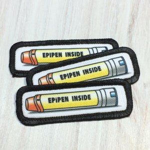 May include: Three white fabric patches with black stitching around the edges. Each patch features a cartoon image of an EpiPen with the text "EPIPEN INSIDE" printed in black.