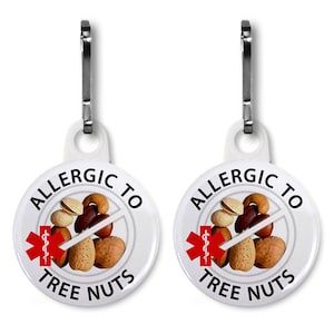 May include: Two white, round medical alert tags with silver-colored clips. Each tag has the words "ALLERGIC TO TREE NUTS" in a circular design, with a red medical symbol and an image of various nuts crossed out.