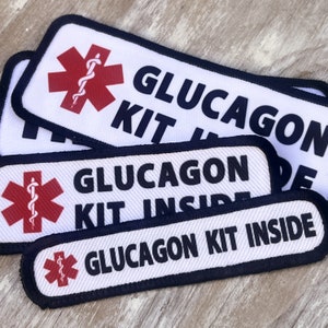 May include: Three white fabric patches with black borders and a red medical symbol. The patches read "Glucagon Kit Inside".