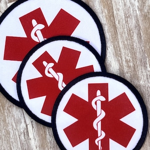 May include: Three white fabric patches with red medical star of life symbols and a snake wrapped around a rod. The patches are different sizes and have black stitching around the edges.