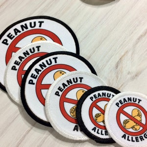 May include: Five white round fabric patches with black borders. Each patch has a red circle with a diagonal line through it and the word "PEANUT" written in black letters. The center of each circle has a graphic of a peanut.