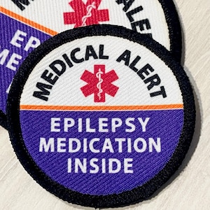 May include: A round, purple and white medical alert patch with black stitching. The patch has a red star of life symbol with a snake wrapped around a rod, and the text "MEDICAL ALERT" in black lettering. Below the star of life is the text "EPILEPSY MEDICATION INSIDE" in white lettering.