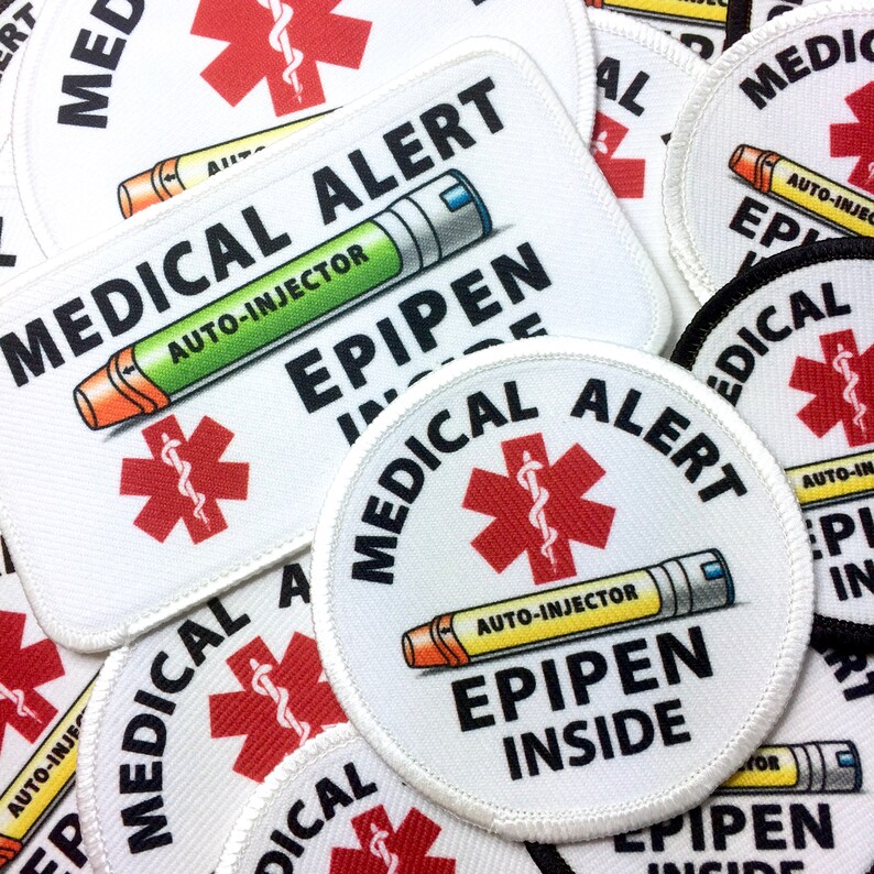 May include: A close-up of several white medical alert patches with a red star of life and the text "MEDICAL ALERT" and "EPIPEN INSIDE". The patches are made of fabric and have a white border.