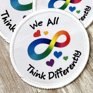 May include: White round fabric patch with a rainbow infinity symbol and the text "We All Think Differently" in black. There are small hearts in different colors around the infinity symbol.