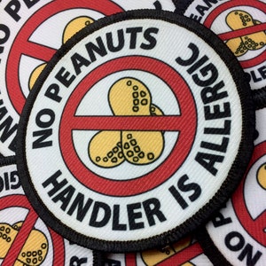 May include: A white round patch with black stitching around the edge. The patch has a red circle with a diagonal red line through it. Inside the circle are three yellow peanut shapes. The text "NO PEANUTS HANDLER IS ALLERGIC" is written in black around the red circle.