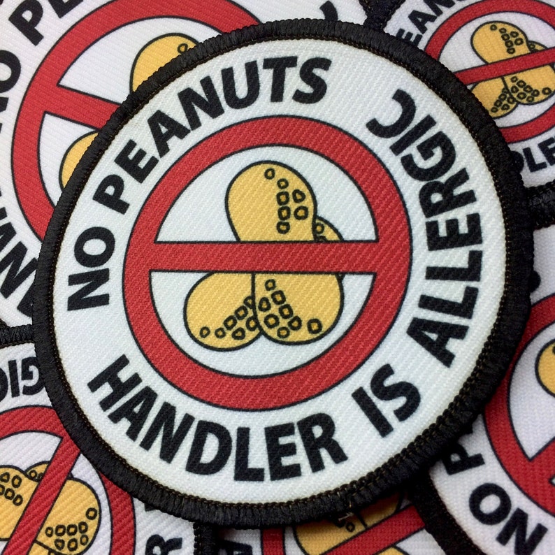 May include: A white round patch with black stitching around the edge. The patch has a red circle with a diagonal red line through it. Inside the circle are three yellow peanut shapes. The text "NO PEANUTS HANDLER IS ALLERGIC" is written in black around the red circle.