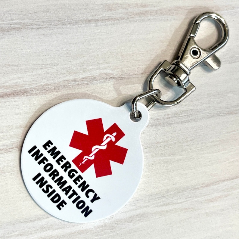 May include: White round keychain with a red star of life symbol and the text "EMERGENCY INFORMATION INSIDE". The keychain has a silver metal clip.