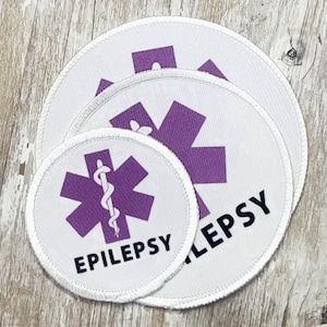 May include: Three white round fabric patches with a purple star of life symbol and the word "EPILEPSY" in black text. The largest patch has the word "EPILEPSY" in a larger font size.