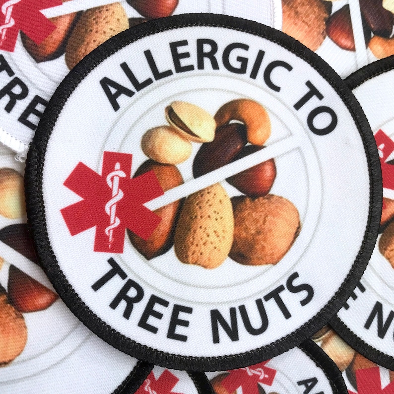May include: A white round patch with black stitching around the edges. The patch has a red medical symbol with a snake wrapped around a staff. The text "Allergic to Tree Nuts" is printed in black around the edge of the patch. A variety of tree nuts are pictured in the center of the patch with a red line drawn through them.