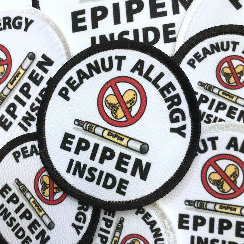 May include: Close-up of several round white patches with black borders. Each patch has a red circle with a diagonal line through it, depicting peanuts. The text "PEANUT ALLERGY" and "EPIPEN INSIDE" is printed in black on each patch.