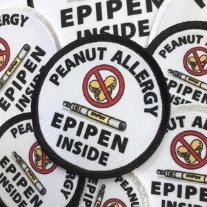 May include: A white round patch with black trim and the text "Peanut Allergy" in black. A red circle with a diagonal line through it contains a drawing of peanuts. A white epipen with the text "EPIPEN" is drawn below the circle. The text "EPIPEN INSIDE" is written in black below the epipen.