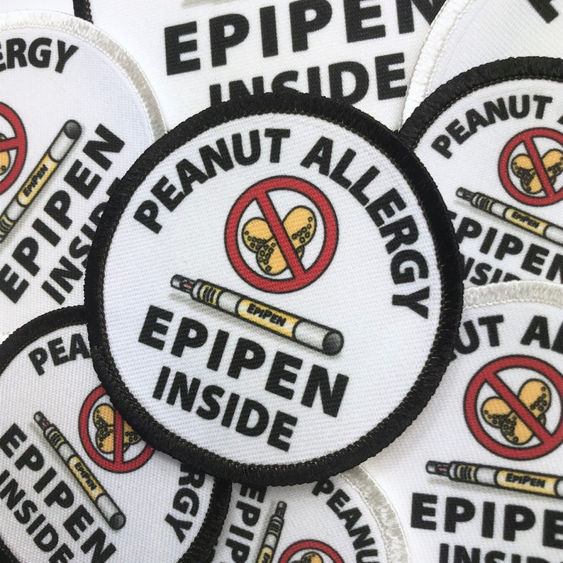 May include: A white round patch with black trim and the text "Peanut Allergy" in black. A red circle with a diagonal line through it contains a drawing of peanuts. A white epipen with the text "EPIPEN" is drawn below the circle. The text "EPIPEN INSIDE" is written in black below the epipen.