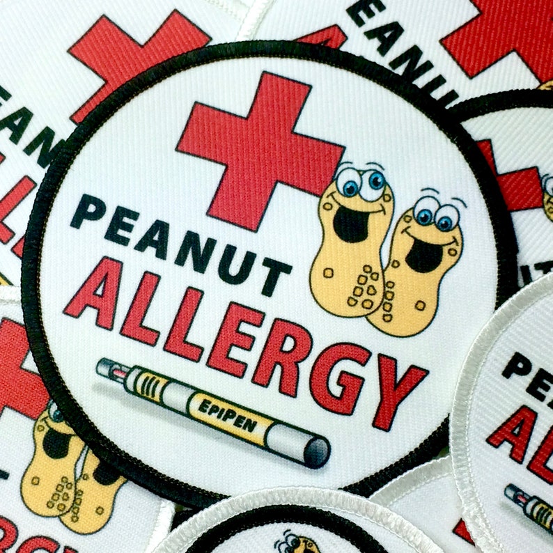 May include: A white round patch with a red cross, the words "Peanut Allergy" in black and red, and a cartoon peanut with a smiling face. The patch also has a red and white epipen with the word "EPIPEN" printed on it.