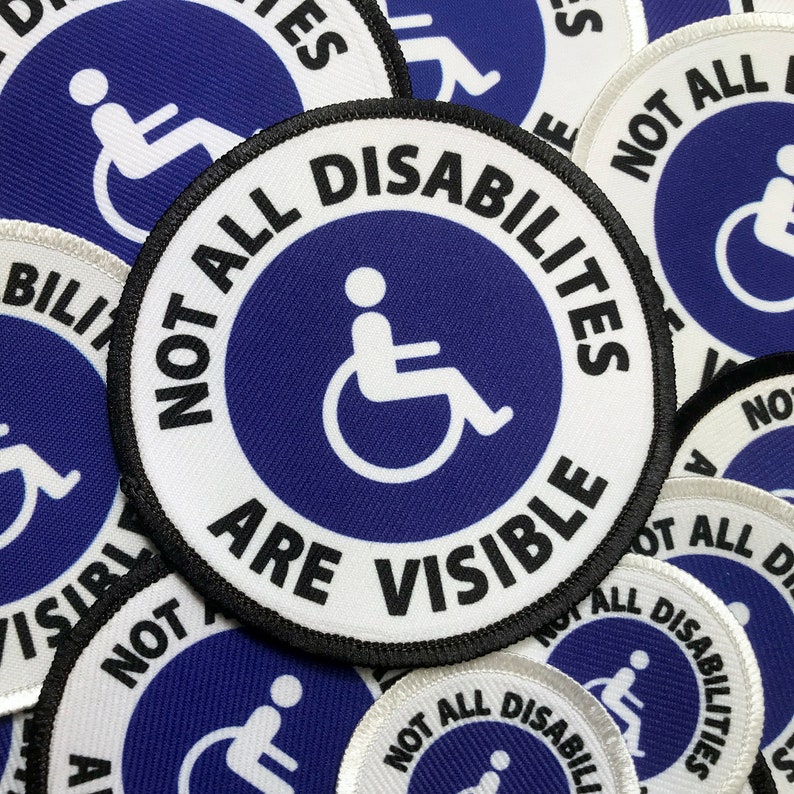 May include: A close-up of several round, blue and white embroidered patches with the text "Not All Disabilities Are Visible" and a white wheelchair symbol in the center. The patches have black stitching around the edges.